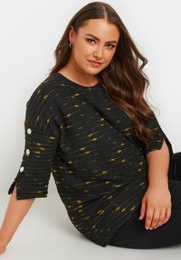 Svart texturerad topp med gula och gröna horisontella ränder, ärmar till armbågen med vita knappdetaljer och en avslappnad passform.