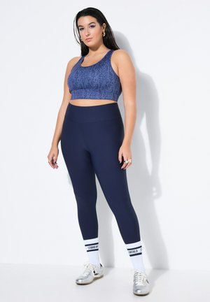 Kvinde stående op ad hvid væg iført blå mønstret sports-bh, marineblå leggings, hvide sokker med sorte striber og sølvfarvede sneakers.