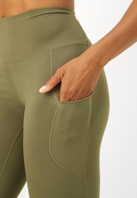 Nära bild av en person som bär olivgröna leggings med hög midja, där en hand vilar innanför en sidoficka på låret.