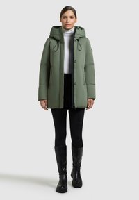 Giacca a vento verde con cappuccio, chiusura lampo frontale e dettagli elasticizzati. Abbinata a un dolcevita bianco e leggings neri, completata con stivali neri.