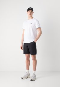 Cămașă albă cu mâneci scurte și accente negre, pantaloni scurți gri și adidași gri. Accesoriile includ o șapcă neagră. Design simplu și croială comodă.