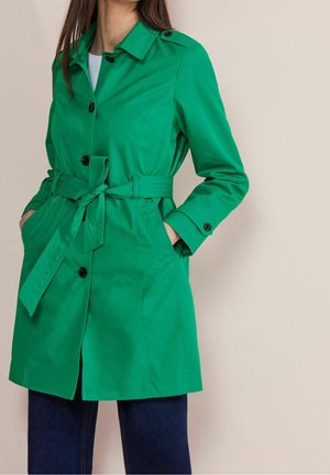 Kobieta ubrana w jaskrawozielony płaszcz trencz z paskiem i czarnymi guzikami, na ciemnych dżinsach, z rękami w kieszeniach, zwrócona lekko w lewo.