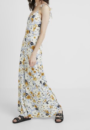Vrouw draagt een lange, mouwloze bloemenjurk met gele, witte en zwarte bloemen, gecombineerd met zwarte open-toe sandalen met zilveren gespen.