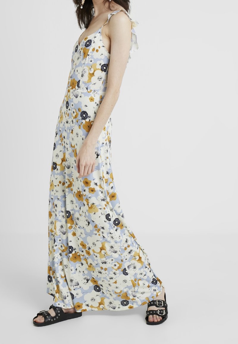Femme portant une longue robe fleurie sans manches avec des fleurs jaunes, blanches et noires, associée à des sandales noires à bout ouvert avec des boucles argentées.