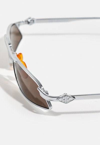 Des lunettes de soleil en métal argenté avec des branches courbées, des verres teintés marron et des coussinets de nez orange, dotées d'un détail de logo en forme de diamant sur la temples.
