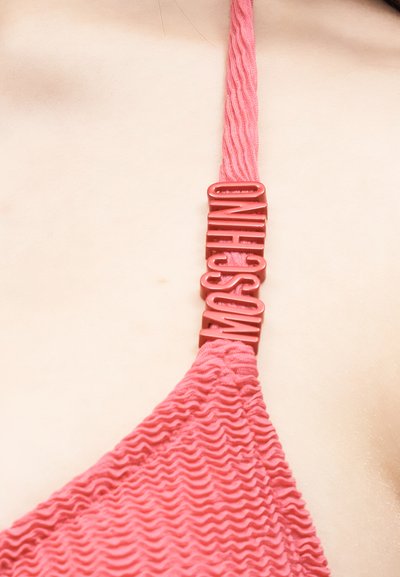 Κοντινό πλάνο ροζ υφασμάτινου λουριού μαγιό με κόκκινα γράμματα που σχηματίζουν τη λέξη "MOSCHINO" στον ώμο και το στήθος ενός ατόμου.