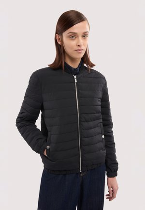 Mujer con chaqueta negra acolchada con cremallera y pantalones oscuros, de pie con una mano en el bolsillo sobre un fondo blanco.