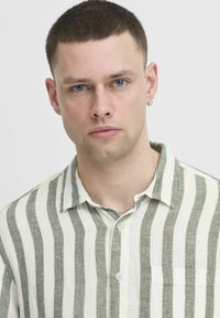 Lněná košile na knoflíky se střídajícími bílými a zelenými vertikálními pruhy, s límečkem a kapsou na hrudi. Hladká textura a lehký materiál.