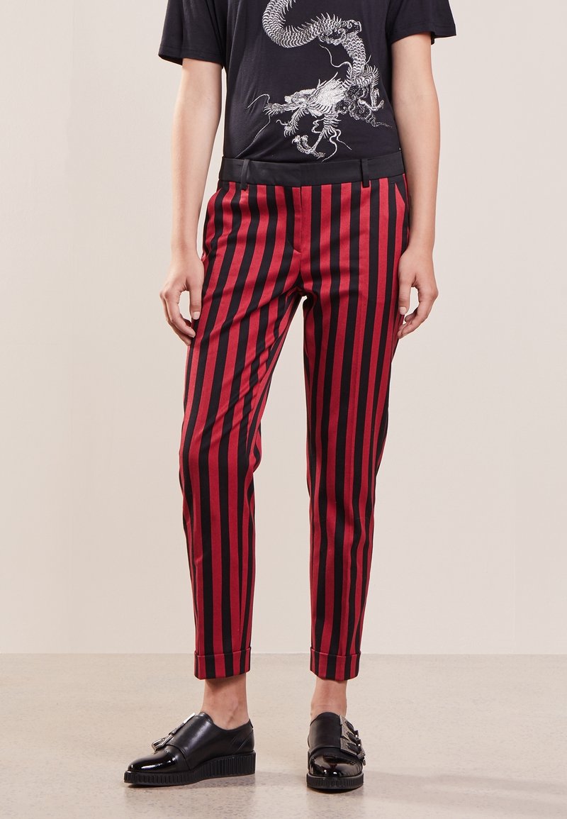 Pantalon rayé rouge et noir avec une coupe slim, présentant une texture lisse, une taille cintrée et des poignets retroussés. Porté avec des chaussures noires brillantes.