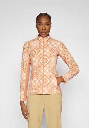 Frau trägt eine zip-up Jacke in Pfirsich- und Cremefarben mit geometrischem Muster und beigefarbene Hosen, steht mit einer Hand in der Tasche vor einfachem Hintergrund.