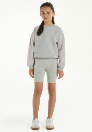 Felpa grigia con maniche a motivo di stelle rosa chiaro, pantaloni corti grigi abbinati. Il modello indossa sneakers bianche con dettagli beige.