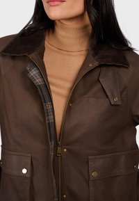 Hnědá kožená bunda s manžetou z kordu, dvěma předními kapsami a kostkovanou podšívkou. Přední zip s měděnými akcenty.