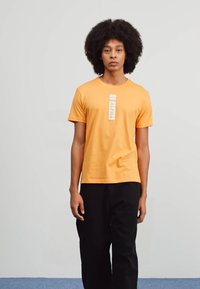 Orange bomuld t-shirt med korte ærmer og rund halsudskæring. Har et hvidt mærke med ordet "ALPHA" centralt på forsiden.