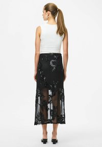 Femme avec queue de cheval portant un haut blanc sans manches et une jupe midi noire transparente à broderies florales, debout de dos sur fond blanc.