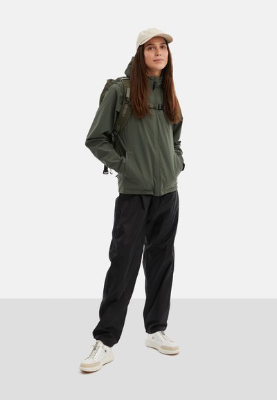 Olivgrön zip-up jacka med huva, svarta löst sittande byxor, krämfärgade sneakers, beige keps och en ryggsäck med justerbara remmar.