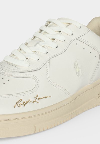 Λευκό δερμάτινο sneaker με μπεζ σόλα, λευκά κορδόνια, χρυσή υπογραφή "Ralph Lauren" και ανάγλυφο λογότυπο Polo στην πλαϊνή πλευρά.