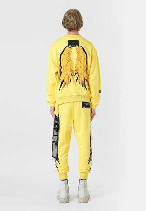 Conjunto de sudadera amarilla y pantalones jogger con diseños de alas en negro y amarillo en la parte posterior, con etiquetas y acentos en los pantalones. Textura suave.
