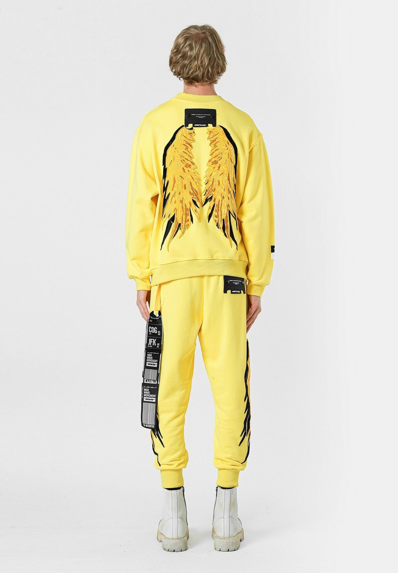 Conjunto de sudadera amarilla y pantalones jogger con diseños de alas en negro y amarillo en la parte posterior, con etiquetas y acentos en los pantalones. Textura suave.