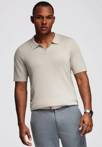 Camisa polo de manga corta beige con cuello plano, puños acanalados y tejido texturizado. Combinada con pantalones de traje gris claro.