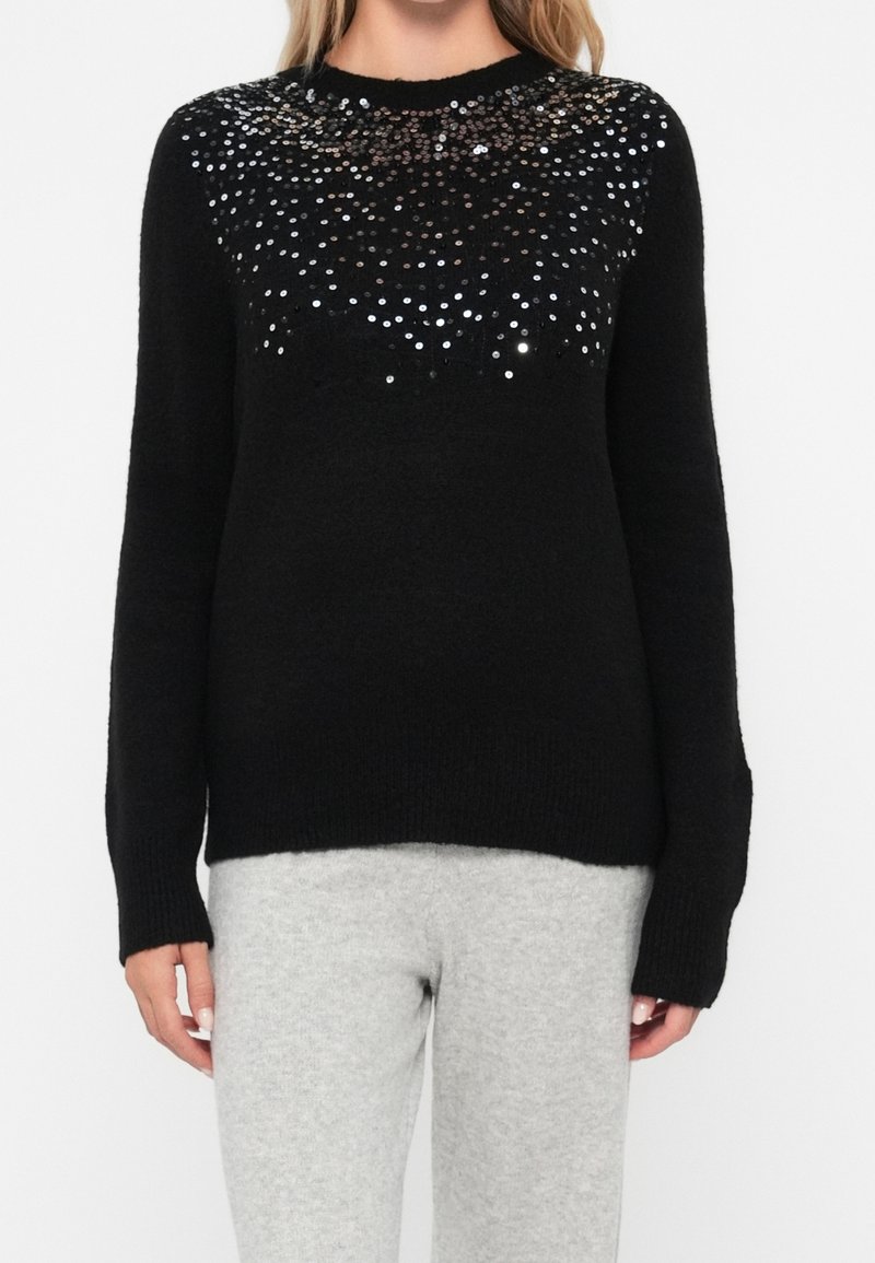 Femme portant un pull en maille noire orné de sequins argentés sur le haut de la poitrine et les épaules, associé à un pantalon gris clair.