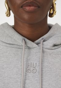 Felpa grigia con cappuccio e cordini, caratterizzata da ricamo "HUGO" in grigio abbinato. Tessuto morbido, design casual e vestibilità rilassata.
