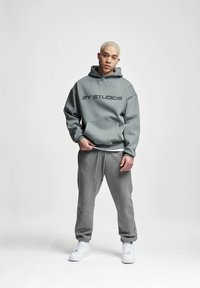 Hoodie grigia con testo "ZY STUDIOS", tasca frontale, vestibilità rilassata; abbinata a pantaloni della tuta grigi e sneaker bianche. Texture liscia.