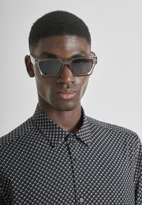 Occhiali da sole con una montatura chiara e angolare e lenti scure, indossati con una camicia a fantasia bianca e nera caratterizzata da piccole forme triangolari.
