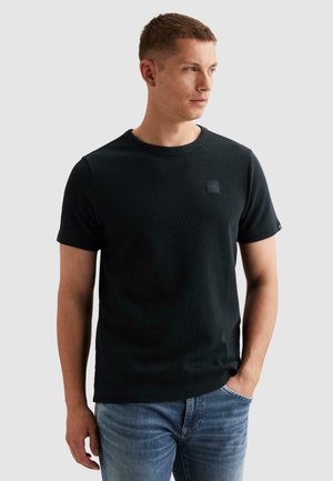 Giovane uomo con t-shirt nera a maniche corte e jeans blu, in piedi con una mano in tasca, che guarda di lato su uno sfondo uniforme.