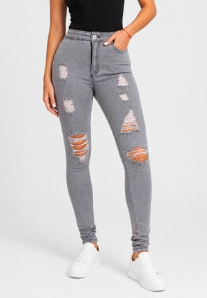 Kvinde iført grå, revet skinny jeans med hvide sneakers og en sort ærmeløs top, stående med den ene hånd i lommen.