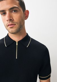 Zwart gebreid poloshirt met een gouden ritssluiting en contrasterende kraag- en mouwboorden. Heeft een gestructureerd patroon en korte mouwen.