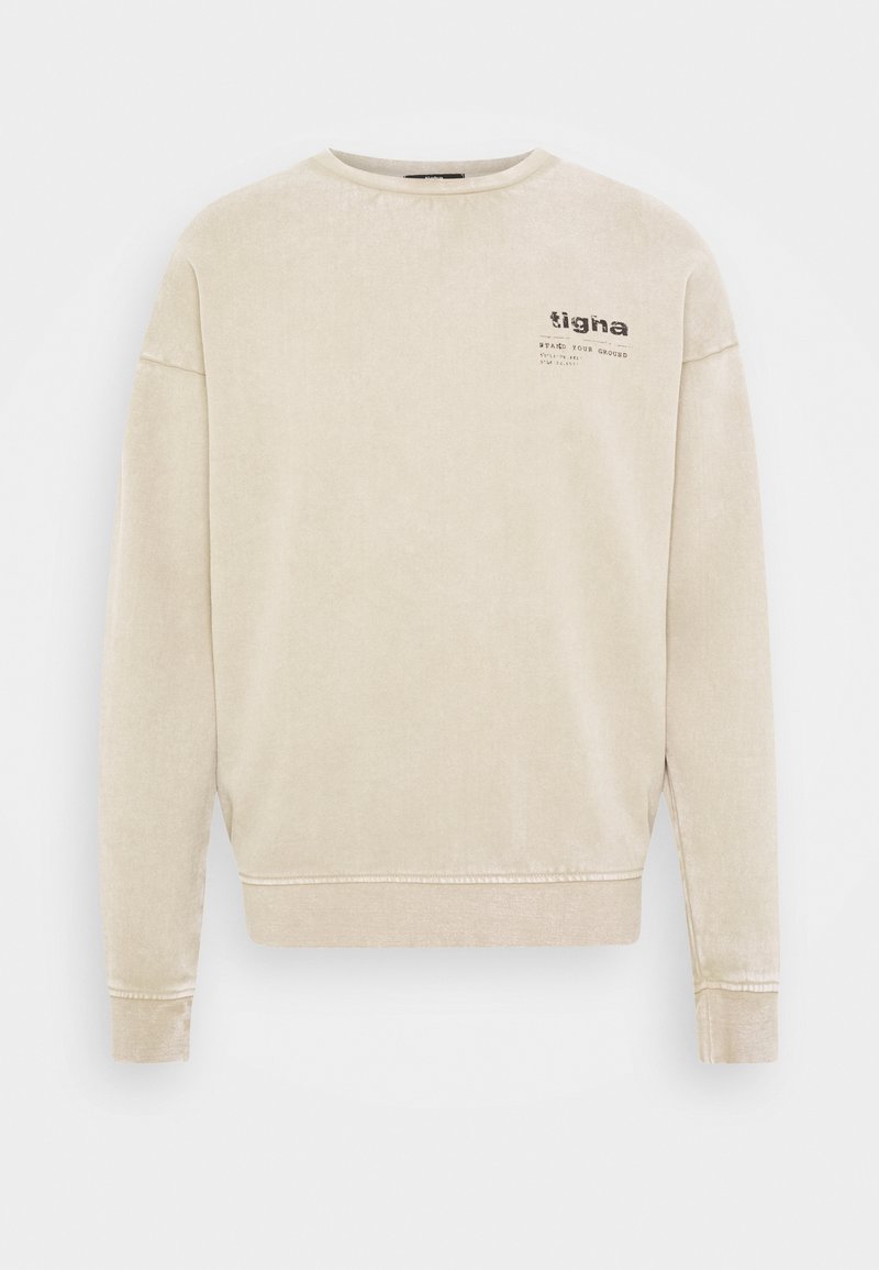 Světlé béžové tričko z měkké látky s kulatým výstřihem, spadlými rukávy a malým grafickým logem na levé horní straně hrudi.