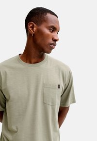 Νεαρός άνδρας με κοντά μαλλιά και μικρό σκουλαρίκι-κρίκο, φοράει ανοιχτό πράσινο μπλουζάκι με στρογγυλό λαιμό και τσέπη στο στήθος, κοιτάζοντας προς τα αριστερά του.