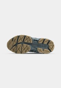 Semelle de chaussure de course avec une bande de roulement en caoutchouc beige et gris foncé comportant des motifs géométriques et des éléments de grip texturés sur fond blanc.
