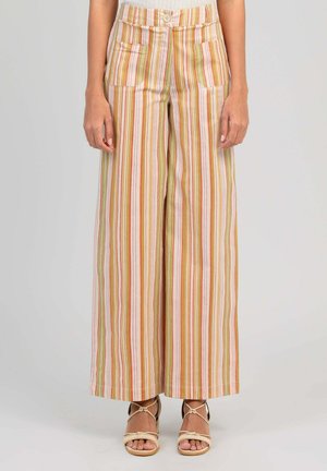 Femme portant un pantalon taille haute à jambes larges avec des rayures verticales orange, vert, blanc et rose, associé à des sandales beiges à brides.
