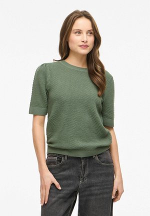 Kvinde med langt brunt hår iført tekstureret grøn kortærmet sweater og sorte jeans, stående med den ene hånd i lommen, kiggende mod højre.