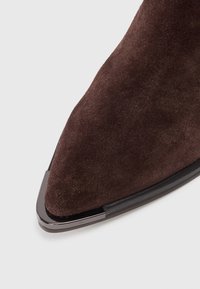 Punta afilada de zapato de ante marrón oscuro con tapa protectora metálica negra en el borde de la suela, presentada sobre fondo blanco.