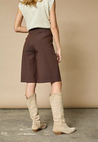 Brune skræddersyede shorts parret med beige ruskind støvler til knæene, der har dekorativ syning. Glat, struktureret stof og afslappet pasform.