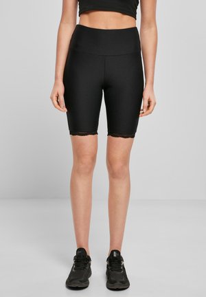Mujer vestida con pantalones cortos biker negros de tiro alto con encaje, top corto negro y zapatillas negras, de pie contra un fondo gris liso.