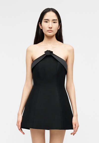 Femme aux longs cheveux noirs portant une mini robe noire bustier avec un détail noué plié en haut, debout devant un fond clair uni.