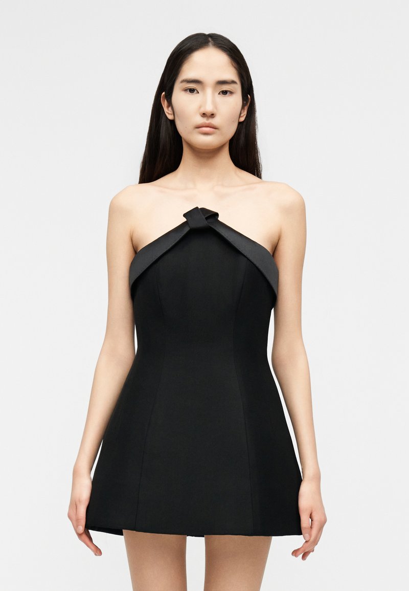 Woman with long dark hair wearing a strapless black mini dress with a folded knot detail at the top, standing against a plain light background.