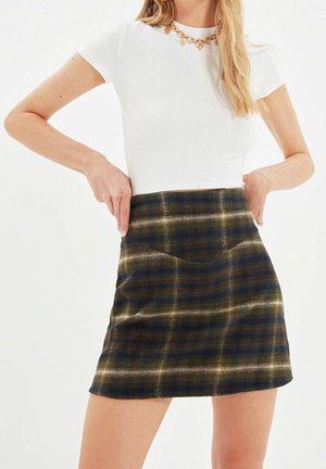 Frau trägt ein weißes Kurzarmtop und einen braun-grünen karierten Minirock, die Taille mit beiden Händen justierend.