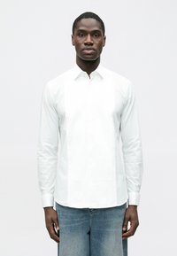 Fehér, hosszú ujjú ing sima anyagból, elülső redőzött panellel, gombos gallérral és mandzsettás ujjakkal. Kék farmerrel viselve.