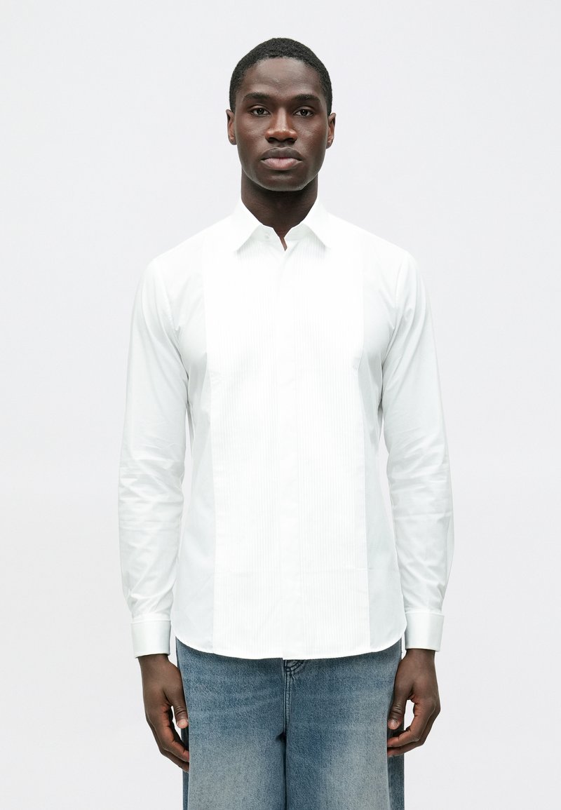 Fehér, hosszú ujjú ing sima anyagból, elülső redőzött panellel, gombos gallérral és mandzsettás ujjakkal. Kék farmerrel viselve.