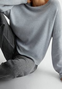 Šedý pletený svetr se širokým výstřihem, s texturovanými akcenty na rukávech, doplněný třpytivými šedými džínami. Volný střih.