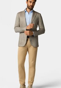 Blazer beige texturé sur une chemise bleu clair, associé à un pantalon kaki et des baskets claires. Les détails notables comprennent une coupe ajustée et des motifs subtils.