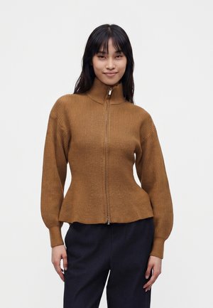 Femme portant un pull marron côtelé zippé avec col montant et manches longues, associé à un pantalon foncé, debout devant un fond uni.