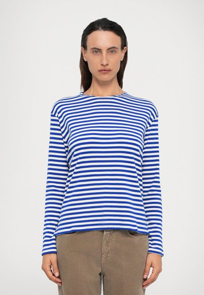 Mujer con cabello oscuro que lleva una camisa de manga larga a rayas azules y blancas y pantalones de pana marrones, de pie contra un fondo blanco liso.