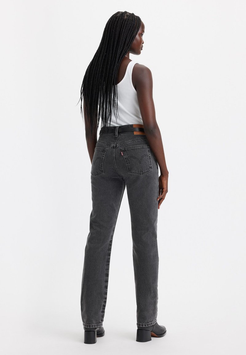 Femme avec de longs cheveux tressés portant un débardeur blanc, un jean noir taille haute et des bottes à talons noires, se tenant contre un fond blanc.