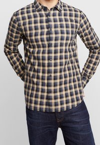 Camicia a maniche lunghe a quadri in beige e blu navy, con colletto button-down, due tasche sul petto e orlo arrotondato. Realizzata in tessuto a trama.