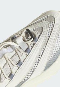 Biela atletická obuv s textúrami z gumy a semišu, s šedými akcentmi, zložitým šitím a systémom šnurovania pre bezpečné uchytenie.